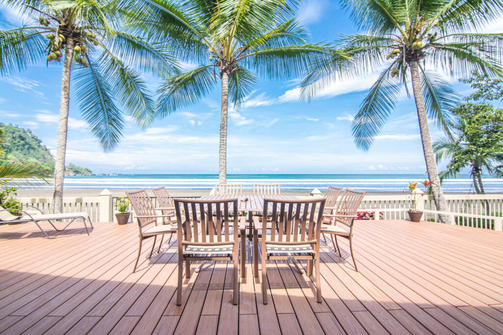 Outdoor dining table and chairs on a beachfront deck framed by coconut palms with ocean views at Casa Rio Mar in Jaco Beach, Costa Rica