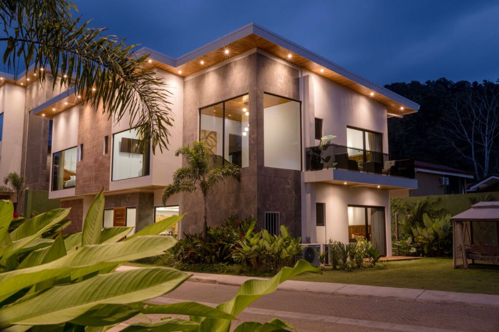 Modern two-storey Casa Rio Mar villa exterior lit at blue hour with floor-to-ceiling windows, balcony and tropical planting in Jaco Beach, Costa Rica