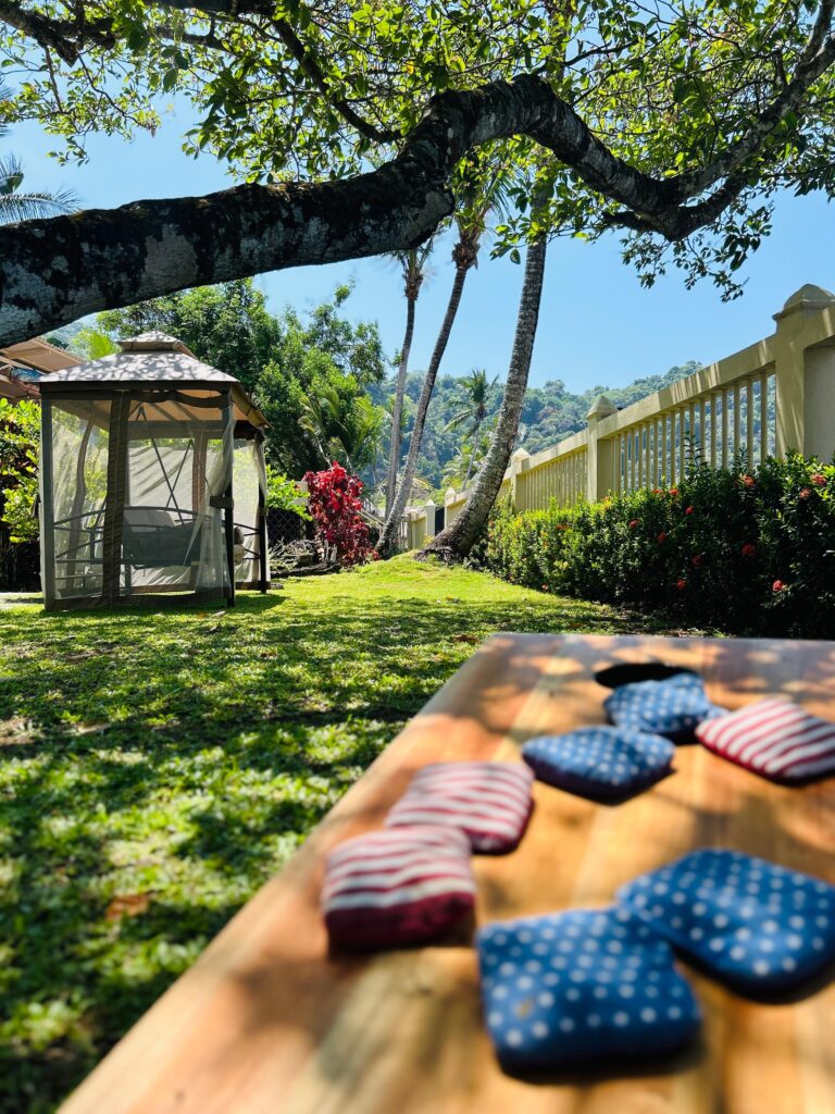 Cornhole board with bean bags set up on the lawn beside a screened gazebo, palm trees and jungle hillside at Casa Rio Mar in Jaco Beach, Costa Rica