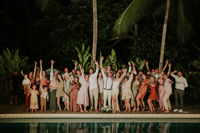 Newlyweds and their wedding guests cheering with raised arms beside the poolside at night, framed by palms and tropical foliage at Casa Rio Mar in Jaco Beach, Costa Rica