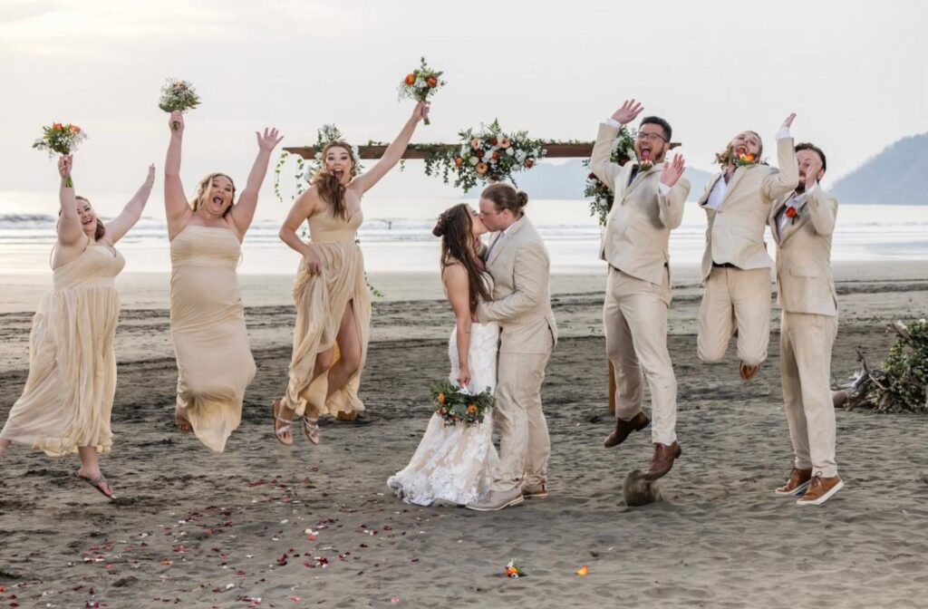 Bride and groom kissing as their wedding party jumps and cheers in neutral attire on the sand at a beach ceremony arch in Jaco Beach, Costa Rica